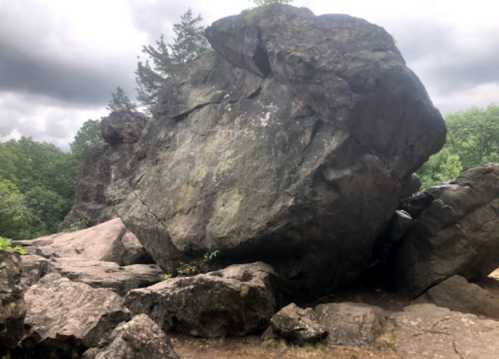 A large, weathered boulder surrounded by smaller rocks and greenery under a cloudy sky.