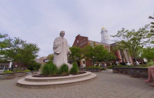 A statue of a robed figure stands in a landscaped area, with a building and trees in the background.