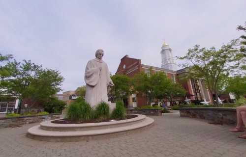 A statue of a robed figure stands in a landscaped area, with a building and trees in the background.