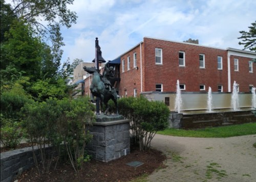 A bronze statue of a horse and rider stands near a fountain in a landscaped area with brick buildings in the background.