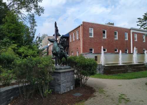 A bronze statue of a horse and rider stands near a fountain in a landscaped area with brick buildings in the background.