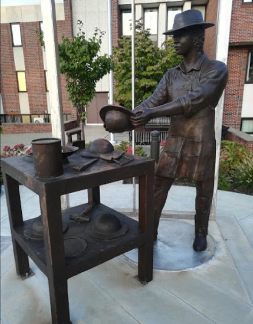 A bronze statue of a person in a hat, preparing food at a table with various cooking pots and utensils.