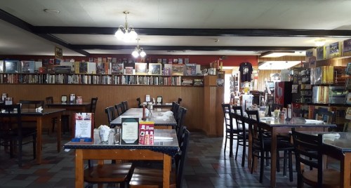 A cozy restaurant interior with wooden tables, chairs, and a bookshelf filled with books along the wall.