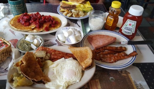 A breakfast spread featuring pancakes with strawberries, eggs, bacon, toast, and sides of syrup and ketchup.