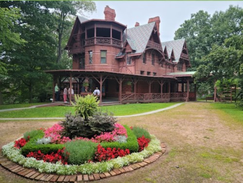 A large, ornate red mansion with a wraparound porch, surrounded by colorful flower beds and lush greenery.
