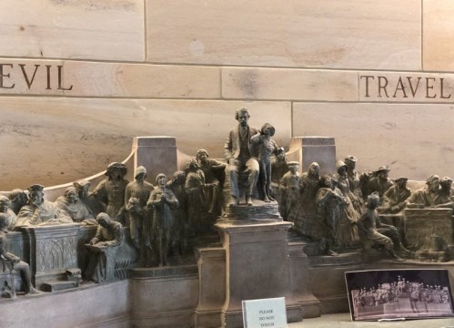 A bronze sculpture depicting historical figures in a gathering, with inscriptions on the wall above.