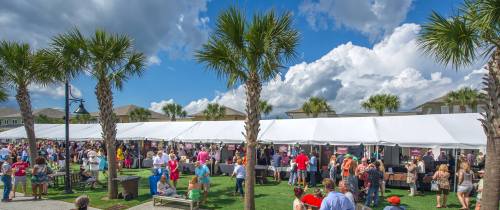 A lively outdoor event with crowds under tents, palm trees, and a bright blue sky with fluffy clouds.