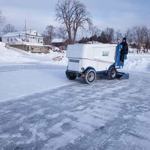 A person operates a Zamboni on an outdoor ice rink, surrounded by snow-covered trees and buildings.