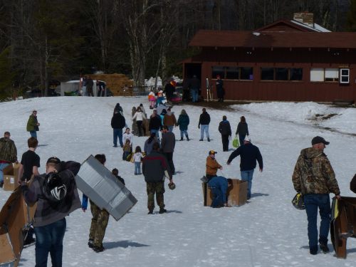 People walking on snow towards a building, carrying boxes and supplies, with trees in the background.