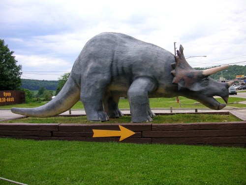 A large, gray dinosaur statue resembling a triceratops, positioned on a grassy area with a directional arrow.