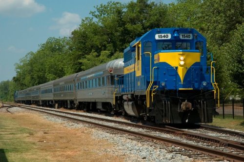 A blue and yellow train engine pulls several silver passenger cars along a tree-lined track on a sunny day.