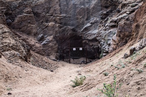 A rocky landscape with a barred entrance to a cave or mine, surrounded by dry earth and sparse vegetation.