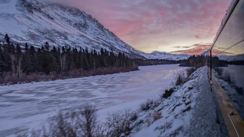 A scenic view of a frozen river and snow-covered mountains at sunset, seen from a train window.