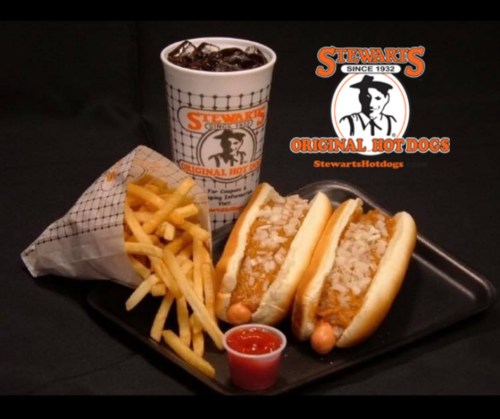 A plate with two hot dogs topped with onions, fries, a cup of soda, and a small container of ketchup.