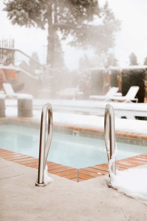 A snowy outdoor pool area with steam rising from the water and lounge chairs in the background.