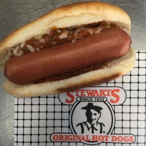 A hot dog in a bun topped with chili and onions, placed on a branded paper with "Stewart's Original Hot Dogs" logo.