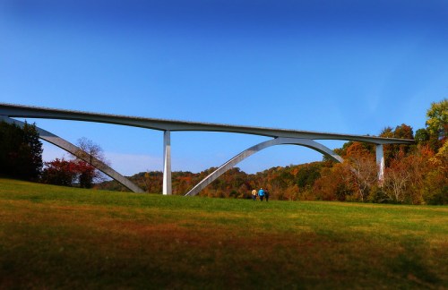 A modern bridge spans a valley, with two people walking on a grassy field beneath it, surrounded by autumn foliage.