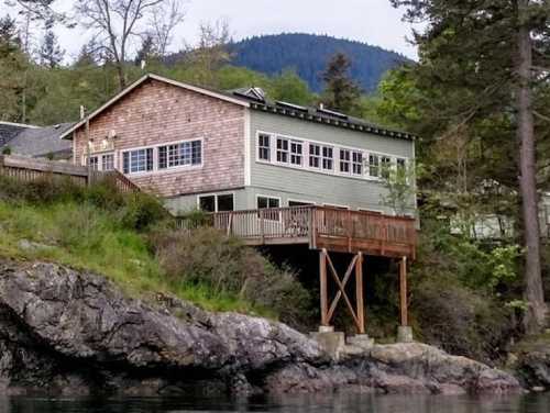 A wooden house on stilts overlooking a rocky shoreline, surrounded by trees and mountains in the background.