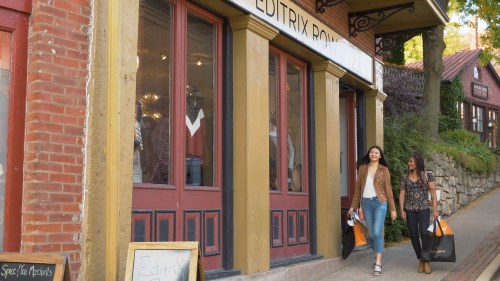Two women walk outside a boutique, carrying shopping bags, with a charming storefront in the background.