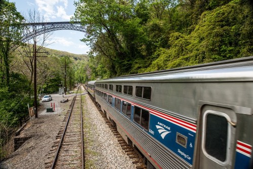 Amtrak train traveling along a scenic route with lush greenery and a bridge in the background.