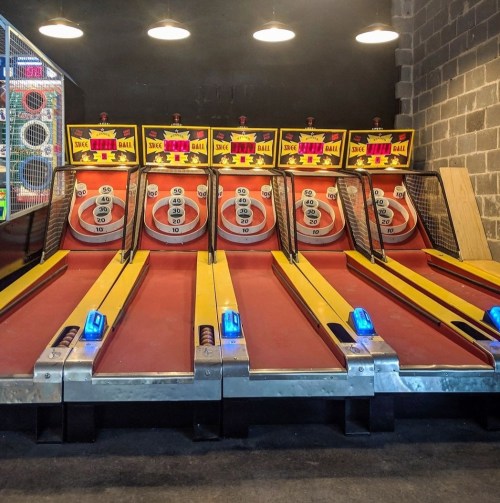 A row of skee ball machines with illuminated scoring areas in a dimly lit arcade.