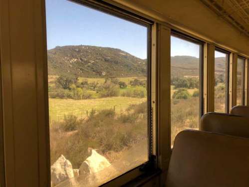 View from a train window showing green fields and rolling hills under a clear blue sky.
