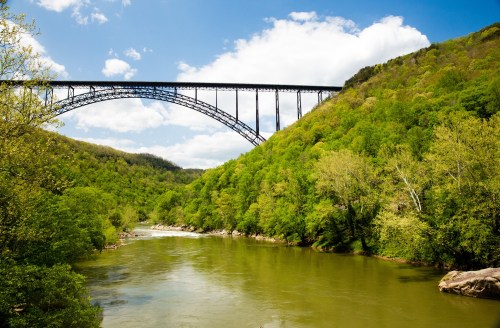 A scenic view of a river surrounded by lush green hills, with a large bridge arching over the landscape under a blue sky.