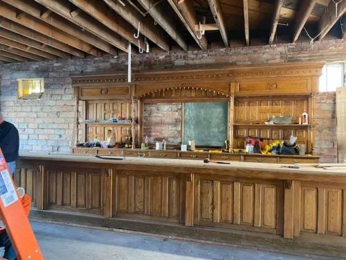 A wooden bar with shelves and a chalkboard, set in a rustic, unfinished space with exposed beams and brick walls.