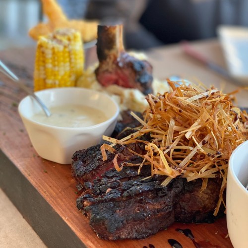 A wooden platter featuring grilled steak, crispy fried onions, corn on the cob, and a small bowl of sauce.