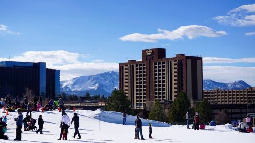 A snowy landscape with people skiing and playing, featuring a hotel and mountains in the background under a clear blue sky.