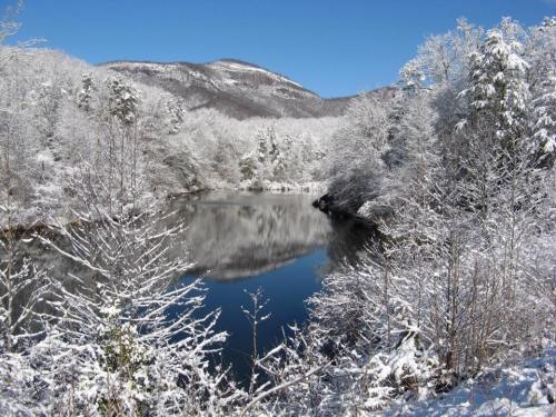 A serene winter landscape featuring a snow-covered river surrounded by trees and mountains under a clear blue sky.