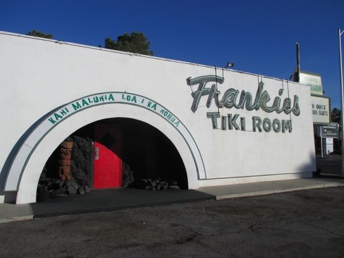 Exterior of Frankie's Tiki Room, featuring a curved entrance, vibrant signage, and a red door against a white wall.