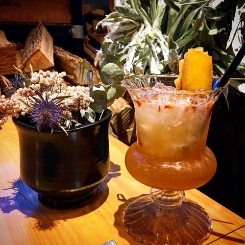 A decorative cocktail in a glass with a garnish, next to a black pot of dried flowers and greenery on a wooden table.