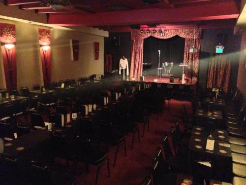 A dimly lit comedy club with empty tables and chairs, featuring a small stage with a microphone and spotlight.