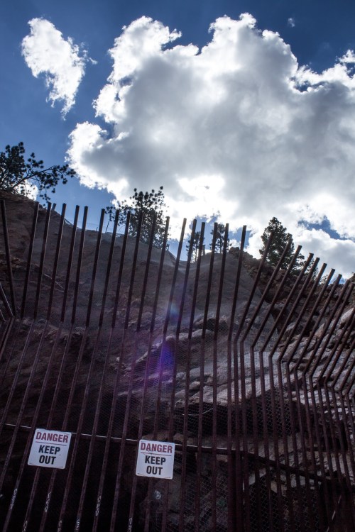 A fence with "Danger Keep Out" signs against a backdrop of clouds and trees under a blue sky.