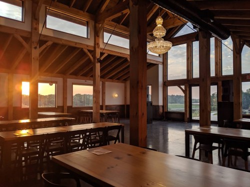 Interior of a rustic restaurant with wooden beams, tables, and large windows showcasing a sunset view.