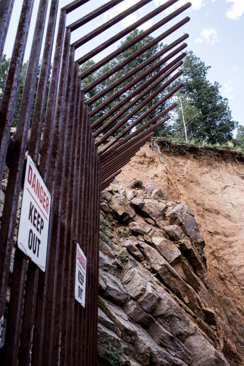 A rusted metal barrier with "Danger" and "Keep Out" signs, next to a steep rocky cliff and trees in the background.