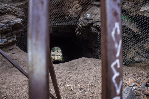 A view through rusty bars into a dark cave entrance, with sandy ground and rocky walls surrounding it.
