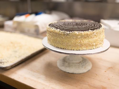 A round cake with chocolate icing and almond flakes, displayed on a white cake stand in a kitchen setting.