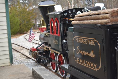A vintage steam locomotive at Heritage Park Railway in Simpsonville, with an American flag and wooden logs on top.