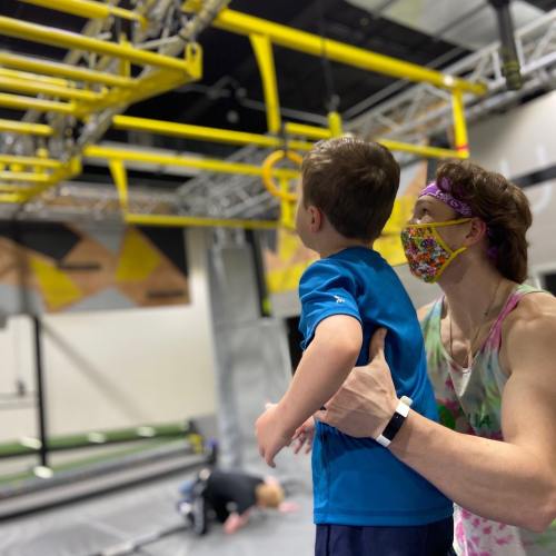 A woman assists a child at a gym, both wearing masks, near climbing equipment and other children in the background.