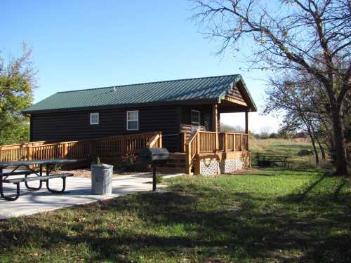 A wooden cabin with a green metal roof, surrounded by grass and trees, featuring a picnic table and a trash can.