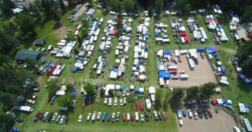 Aerial view of a large outdoor event with numerous tents, vehicles, and people in a green park setting.