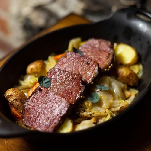 A cast iron skillet filled with sliced beef, potatoes, cabbage, and carrots, garnished with fresh herbs.