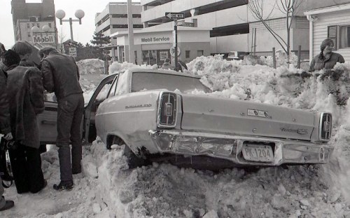 A vintage car stuck in deep snow, with people nearby assessing the situation in a wintery urban setting.