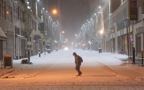 A lone figure walks through a snow-covered, deserted street during a snowstorm, with buildings and streetlights in the background.