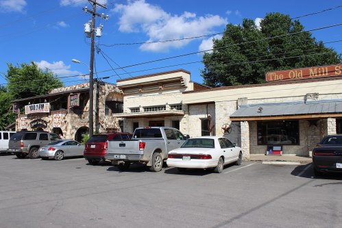 A row of buildings with shops, parked cars in front, under a blue sky with scattered clouds.