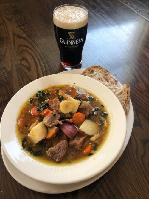 A bowl of hearty beef stew with vegetables and potatoes, served with a slice of bread and a pint of Guinness.
