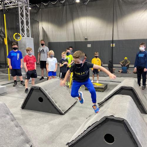 A group of children in masks watches as one child jumps over an obstacle in a ninja training gym.