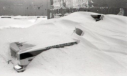 A snow-covered truck partially buried in deep snow, with only the top visible, against a brick wall.